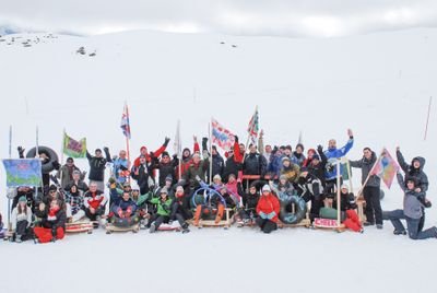 Gruppe von Menschen im Schnee mit Schlitten und Fahnen, umgeben von verschneiten Bergen.