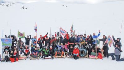 Gruppe von Menschen im Schnee mit Schlitten und Fahnen, umgeben von verschneiten Bergen.