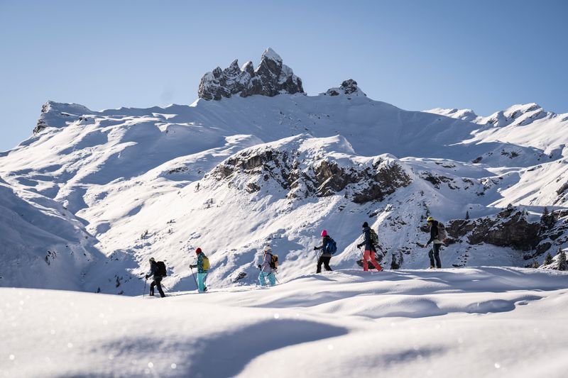 Personen mit Skiausrüstung wandern durch verschneite Berglandschaft unter klarem Himmel.