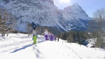 Personen in bunter Winterkleidung wandern im Schnee vor einer Bergkulisse.