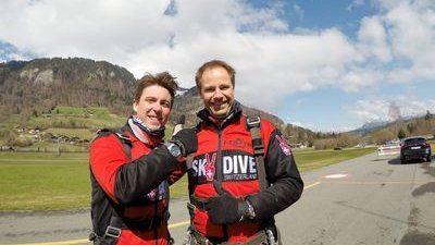 Zwei Personen in roten Fallschirmspringeranzügen und Gurten stehen auf einem Flugplatz vor Bergen.
