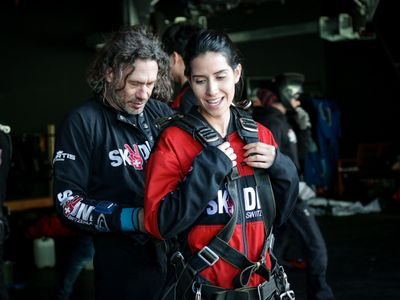 Personen mit Fallschirmgurten in einem Innenraum, eine Person hilft der anderen beim Anlegen der Ausrüstung.