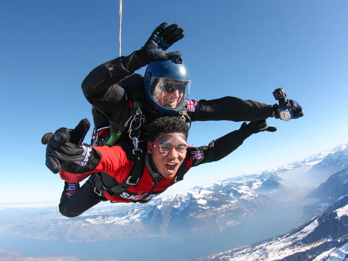 Two people tandem skydiving over snow-covered mountains, wearing helmets and goggles.