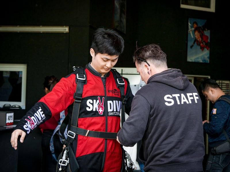 Person in skydiving suit is being prepared by employee with safety harness.