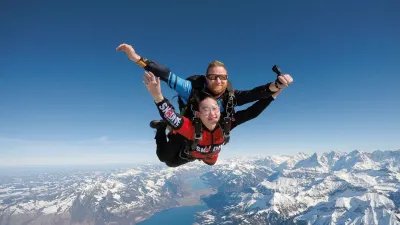 Two people tandem skydiving with helmets and suits over snowy Alps.