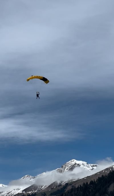 Person with a yellow parachute is floating over snow-covered mountains under a blue sky.