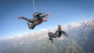 Fallschirmspringer in der Luft mit Helm und Anzug über Berglandschaft, begleitet von einem Guide.