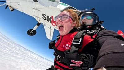 Zwei Personen beim Tandem-Fallschirmsprung mit Helmen und Schutzbrillen über einer Wolkendecke.