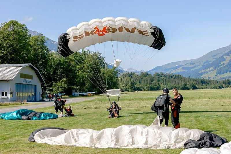 Fallschirmspringer landen auf einer Wiese vor einem Hangar in den Schweizer Alpen, mit Fallschirmen.