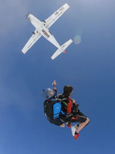 Zwei Personen im Tandem-Fallschirmsprung mit Helmen und Gurten, Flugzeug im Hintergrund, blauer Himmel.