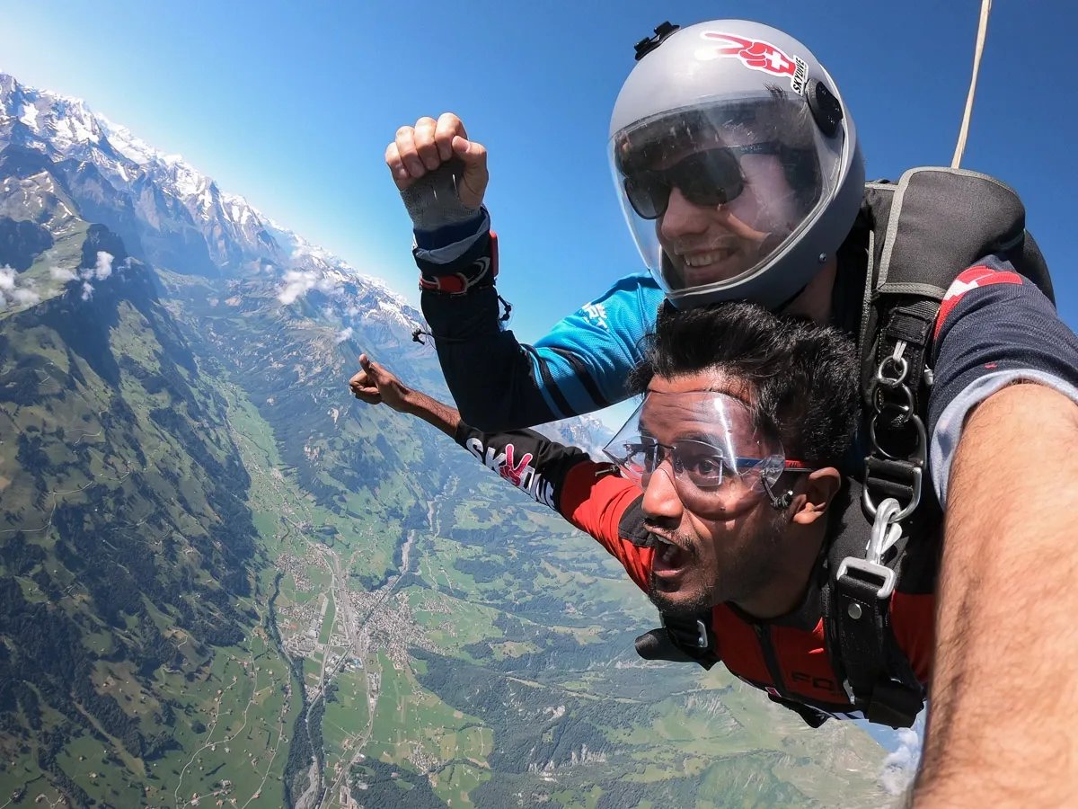 Two people tandem skydiving over a mountain landscape, wearing helmets and goggles.