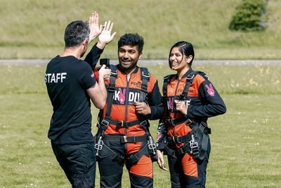 Zwei Personen in Fallschirmspringeranzügen mit Gurten auf einer Wiese, begleitet von einem Mitarbeiter.
