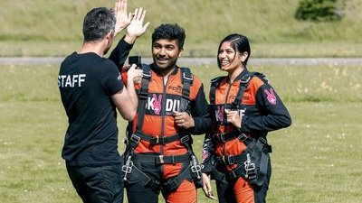 Zwei Personen in Fallschirmspringeranzügen mit Gurten auf einer Wiese, begleitet von einem Mitarbeiter.